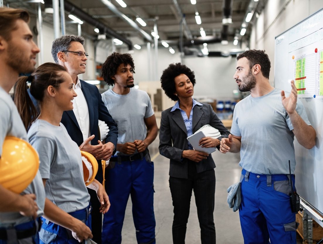 A group of workers in industrial setting discussing a project in front of a white board
