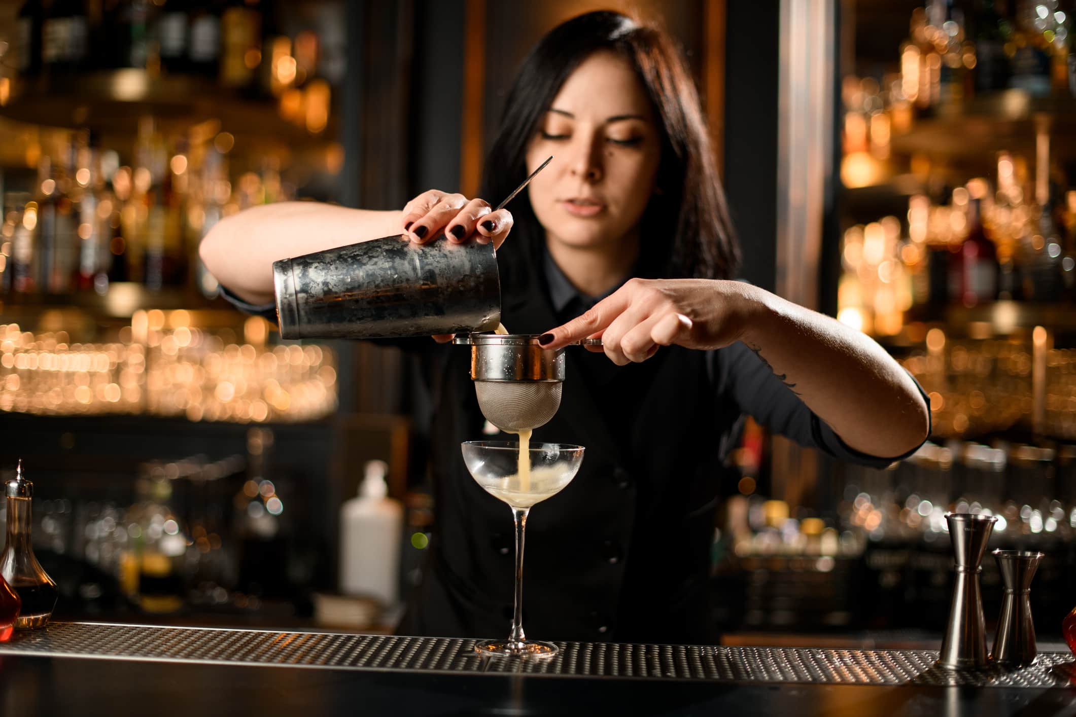 Cute female bartender makes cocktail drink with a steel sieve
