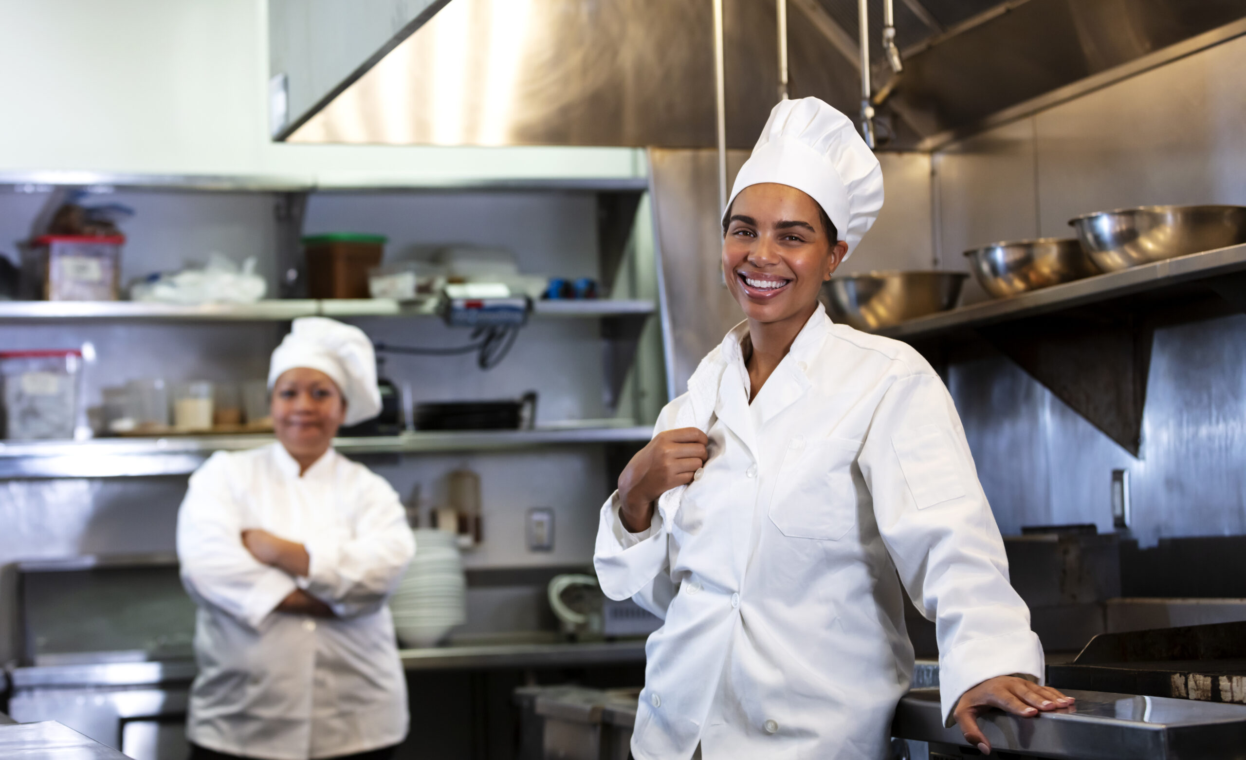 Two women in their 30s working as chefs in a restaurant. They are in the commercial kitchen wearing chef's whites, looking over the service counter.